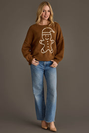 A person wearing a Brown Gingerbread Sweater featuring a white gingerbread man design and blue wide-leg jeans.