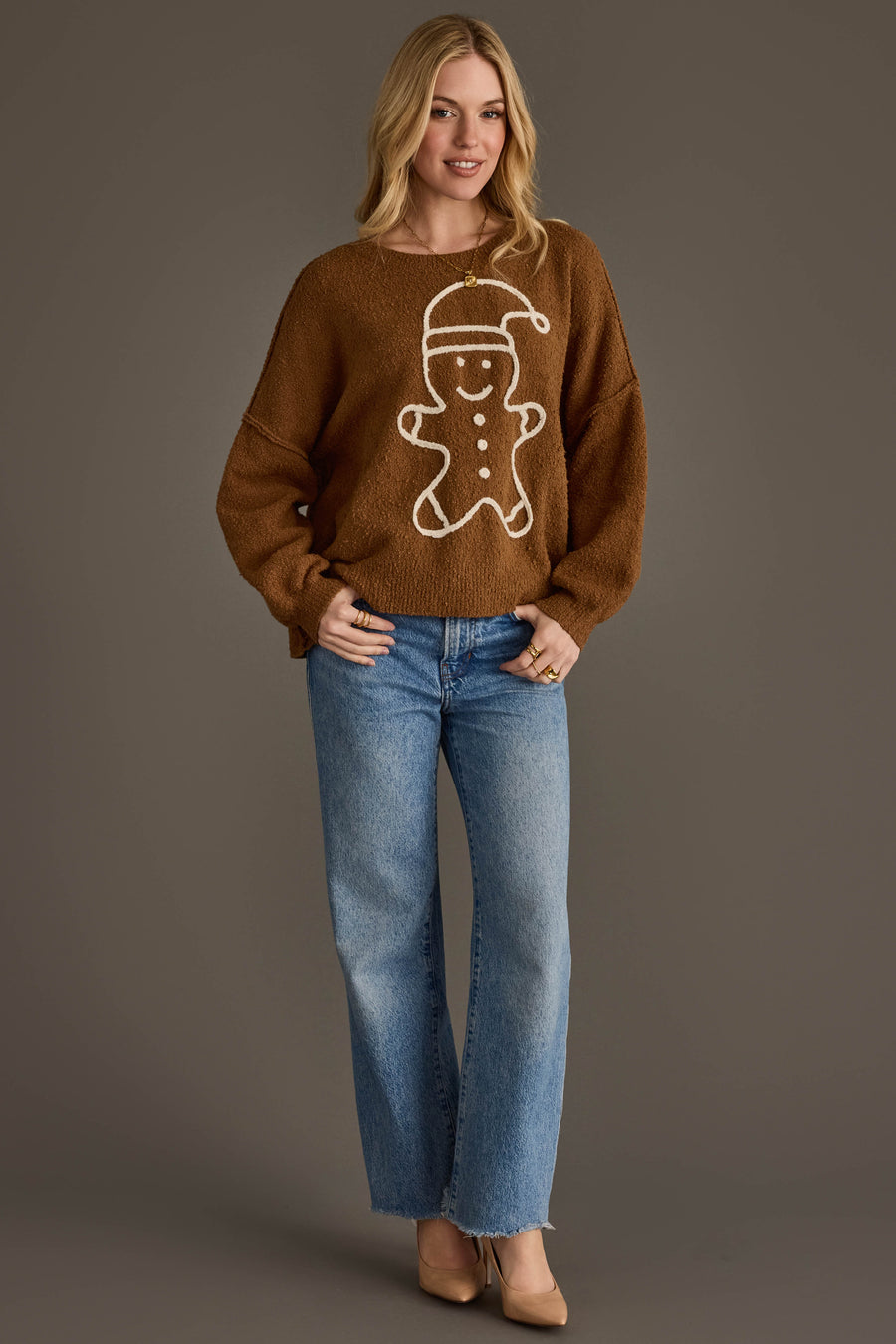 A person wearing a Brown Gingerbread Sweater featuring a white gingerbread man design and blue wide-leg jeans.
