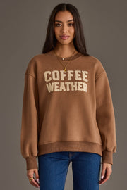 A woman with long dark hair models the Panache Apparel Collection Light Brown Coffee Weather Sweatshirt, featuring cream lettering, paired with layered necklaces and blue jeans, standing against a plain gray background.