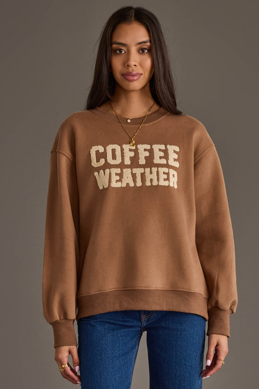 A woman with long dark hair models the Panache Apparel Collection Light Brown Coffee Weather Sweatshirt, featuring cream lettering, paired with layered necklaces and blue jeans, standing against a plain gray background.