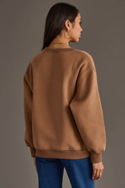 A woman with long dark hair wears gold hoop earrings, blue jeans, and the Light Brown Coffee Weather Sweatshirt from Panache Apparel Collection. She is standing with her back to the camera against a plain background.
