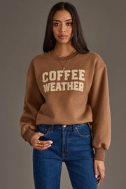 A woman with long dark hair is wearing the Panache Apparel Collection’s Light Brown Coffee Weather Sweatshirt and blue jeans, standing against a plain gray background.