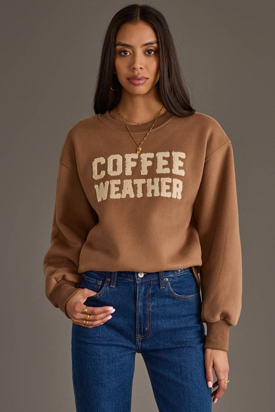 A woman with long dark hair is wearing the Panache Apparel Collection’s Light Brown Coffee Weather Sweatshirt and blue jeans, standing against a plain gray background.