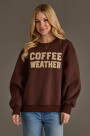 A woman with long blonde hair wears the Panache Apparel Collection Dark Brown Coffee Weather Sweatshirt, featuring beige lettering, paired with blue jeans while standing against a plain gray background.