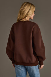 A person with long blonde hair, seen from behind, is wearing the Panache Apparel Collection’s Dark Brown Coffee Weather Sweatshirt and blue jeans against a plain gray background.