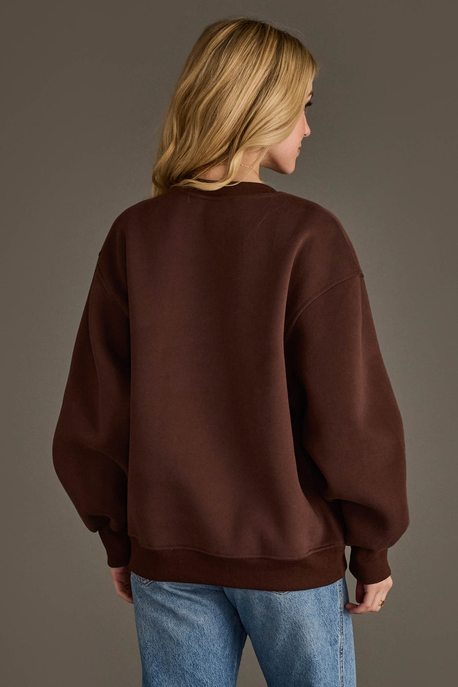 A person with long blonde hair, seen from behind, is wearing the Panache Apparel Collection’s Dark Brown Coffee Weather Sweatshirt and blue jeans against a plain gray background.