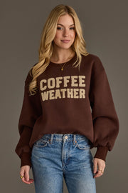 A woman with long blonde hair wears the Panache Apparel Collection Dark Brown Coffee Weather Sweatshirt paired with blue jeans. She stands against a plain gray background, smiling slightly.