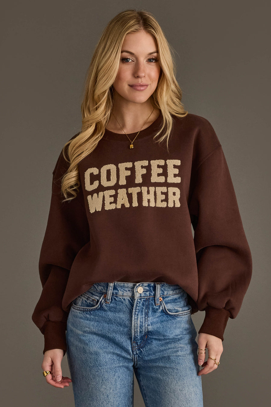 A woman with long blonde hair wears the Panache Apparel Collection Dark Brown Coffee Weather Sweatshirt paired with blue jeans. She stands against a plain gray background, smiling slightly.