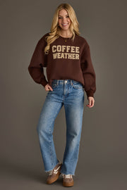 A smiling woman stands against a gray background, wearing the Panache Apparel Collection Dark Brown Coffee Weather Sweatshirt, blue jeans, brown sneakers, and gold necklaces. She has long blonde hair and her hands are relaxed at her sides.