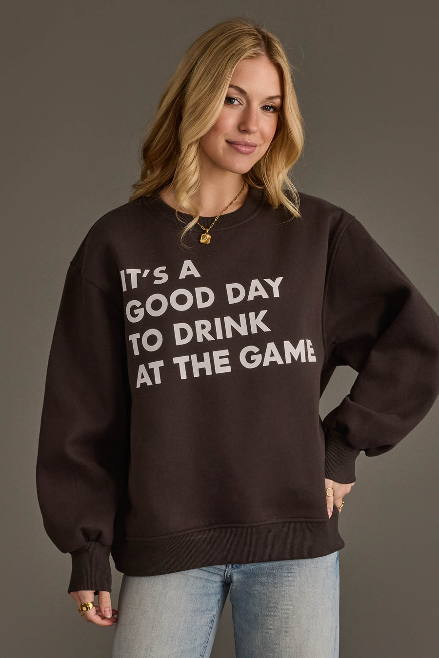 A blonde-haired woman poses against a plain gray background, wearing the Panache Apparel Collection It's A Good Day To Drink At The Game Sweatshirt and placing one hand on her hip.