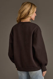 A woman with blonde hair stands facing away, wearing the Panache Apparel Collection "It's A Good Day To Drink At The Game" sweatshirt in dark brown and light blue jeans, against a plain gray background.