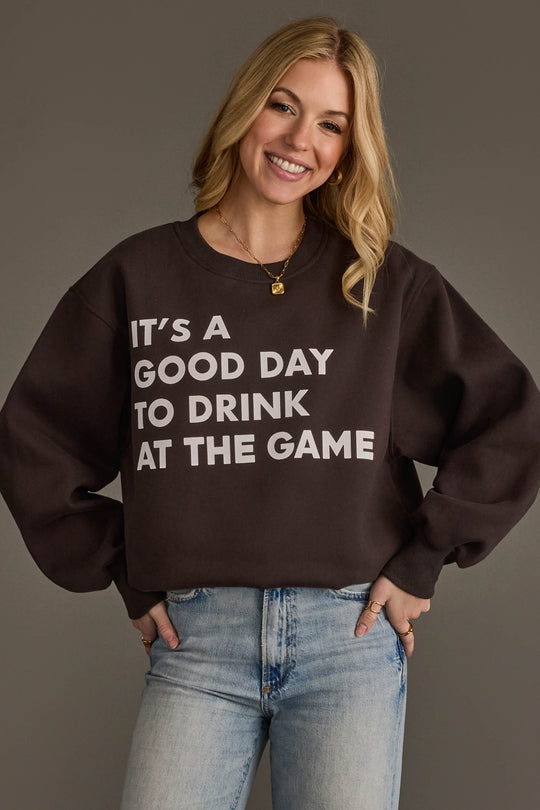A smiling woman with long blonde hair wears the Panache Apparel Collection's "It's A Good Day To Drink At The Game Sweatshirt" paired with light blue jeans, standing against a gray background.