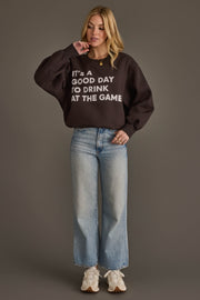 A woman with blonde hair stands against a gray background, touching her chin, wearing the Panache Apparel Collection's "It's A Good Day To Drink At The Game" black sweatshirt, paired with light blue wide-leg jeans and white sneakers.
