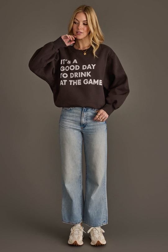A woman with blonde hair stands against a gray background, touching her chin, wearing the Panache Apparel Collection's "It's A Good Day To Drink At The Game" black sweatshirt, paired with light blue wide-leg jeans and white sneakers.