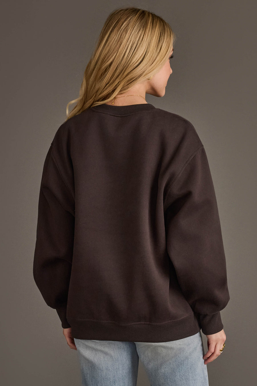 A person with long blonde hair, seen from behind, is wearing the Panache Apparel Collection's "Sorry Your Team Sucks" sweatshirt in a loose dark brown fit and light blue jeans against a neutral background.