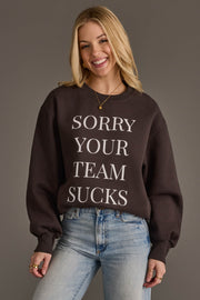 A smiling person with long blonde hair is wearing the Panache Apparel Collection's Sorry Your Team Sucks Sweatshirt—a black sweatshirt with bold white lettering—paired with light blue jeans and standing against a plain background.