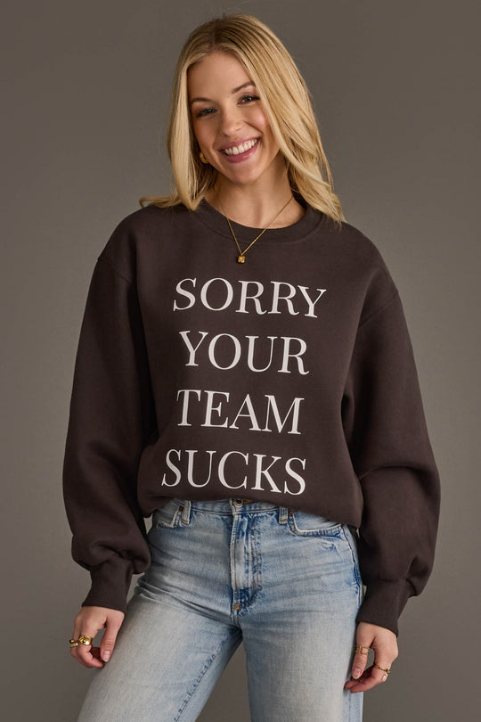 A smiling person with long blonde hair is wearing the Panache Apparel Collection's Sorry Your Team Sucks Sweatshirt—a black sweatshirt with bold white lettering—paired with light blue jeans and standing against a plain background.