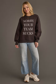 A woman stands smiling against a plain gray background, wearing the Panache Apparel Collection "Sorry Your Team Sucks" Sweatshirt in black, paired with light blue jeans and white sneakers.