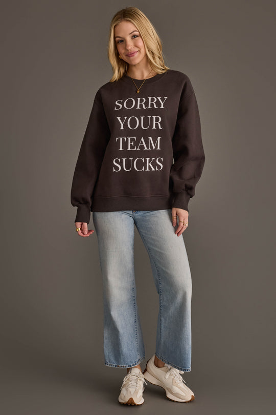 A woman stands smiling against a plain gray background, wearing the Panache Apparel Collection "Sorry Your Team Sucks" Sweatshirt in black, paired with light blue jeans and white sneakers.