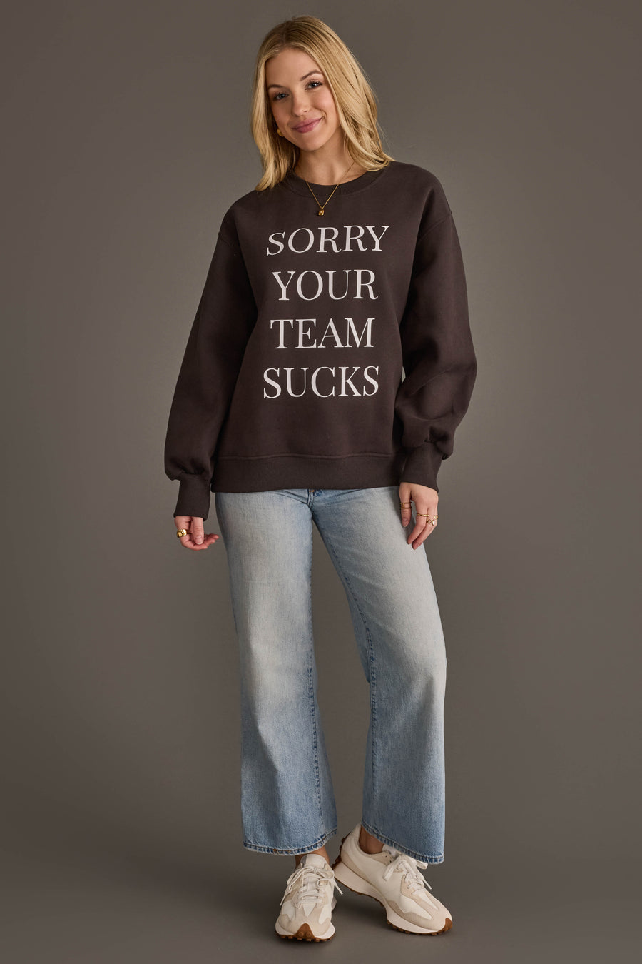 A woman stands smiling against a plain gray background, wearing the Panache Apparel Collection "Sorry Your Team Sucks" Sweatshirt in black, paired with light blue jeans and white sneakers.