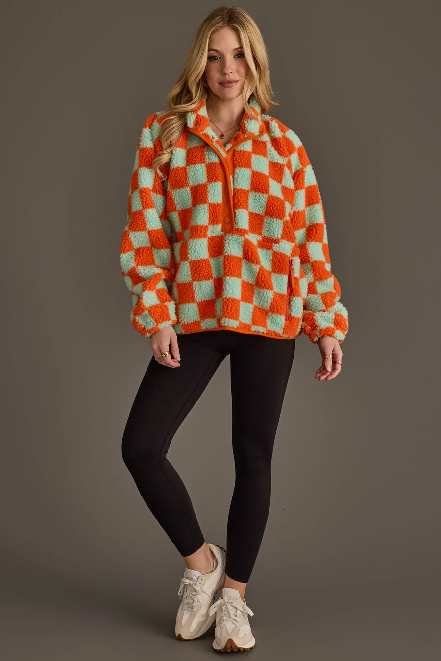 A woman stands against a plain brown background wearing black leggings, cream sneakers, and the Panache Apparel Collection Orange & Light Blue Checkered Fleece Sweatshirt.