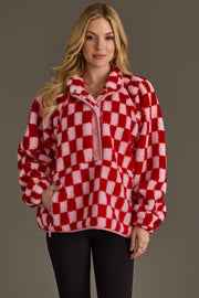 A woman with long blond hair stands against a plain gray background, looking at the camera with a slight smile while wearing the Panache Apparel Collection Light Pink & Red Checkered Fleece Sweatshirt and black pants.