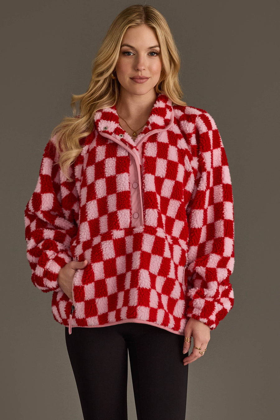 A woman with long blond hair stands against a plain gray background, looking at the camera with a slight smile while wearing the Panache Apparel Collection Light Pink & Red Checkered Fleece Sweatshirt and black pants.