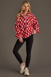 A person wearing the Light Pink & Red Checkered Fleece Sweatshirt from Panache Apparel Collection, paired with black leggings and white sneakers, stands against a plain gray background, smiling with one hand in their pocket.