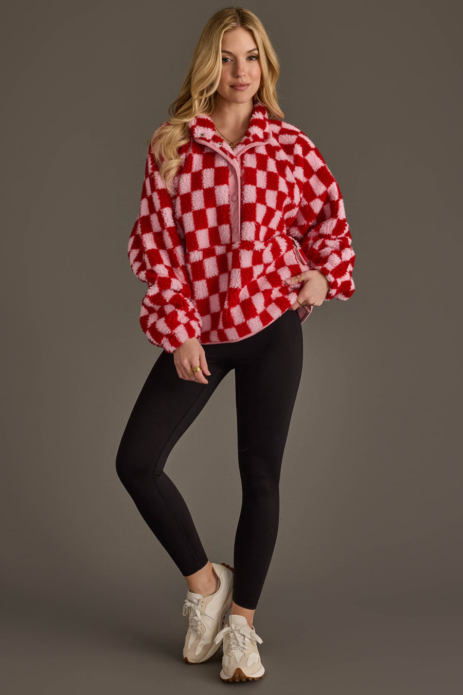 A person wearing the Light Pink & Red Checkered Fleece Sweatshirt from Panache Apparel Collection, paired with black leggings and white sneakers, stands against a plain gray background, smiling with one hand in their pocket.