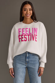 A smiling person wearing a cozy white Feelin Festive Sweatshirt from the Panache Apparel Collection with the pink text FEELIN FESTIVE on the chest and blue jeans.