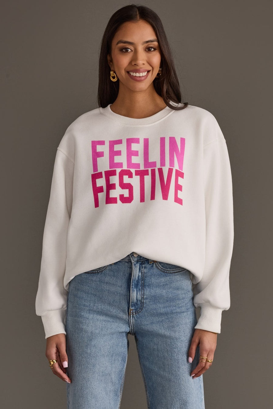 A smiling person wearing a cozy white Feelin Festive Sweatshirt from the Panache Apparel Collection with the pink text FEELIN FESTIVE on the chest and blue jeans.