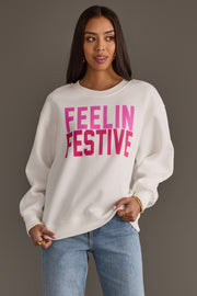 An oversized white Feelin Festive Sweatshirt featuring the words FEELIN FESTIVE in pink block lettering.