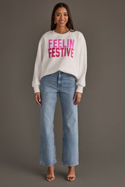 A woman wearing a white Feelin Festive Sweatshirt with pink lettering and light-wash blue jeans.