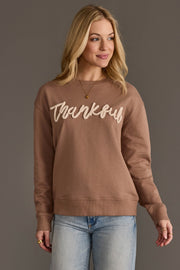 A woman with blonde hair stands against a gray background, smiling slightly as she looks off to her left. She is wearing the Panache Apparel Collection Brown Thankful Sweatshirt, featuring “Thankful” in script across the front, paired with light blue jeans.