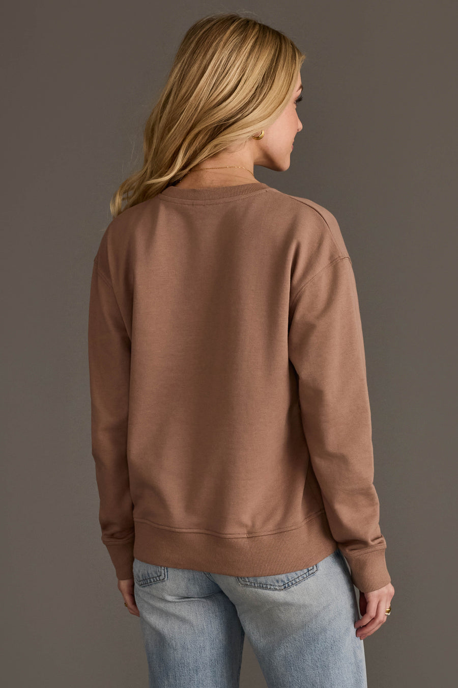 A person with long blonde hair, wearing the Panache Apparel Collection’s Brown Thankful Sweatshirt and light blue jeans, stands facing away against a plain grey background.