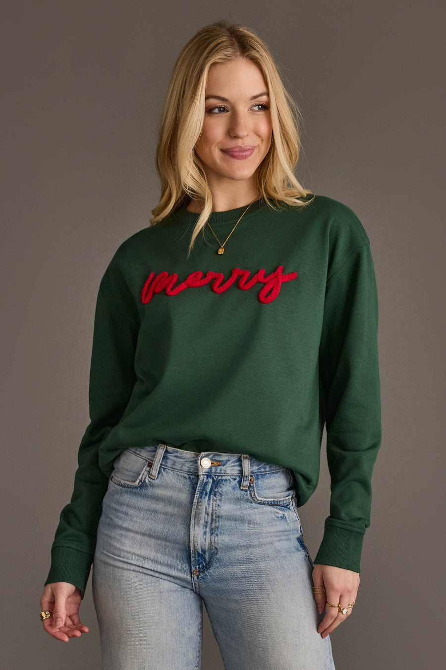 Woman wearing a dark green Dark Green Merry Sweatshirt with the word merry in red script embroidery on the chest.