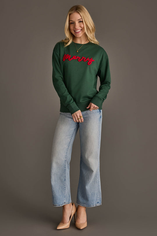 A person wearing a Dark Green Merry Sweatshirt with merry embroidered in red script across the chest, paired with light-wash wide-leg jeans and tan high heels.