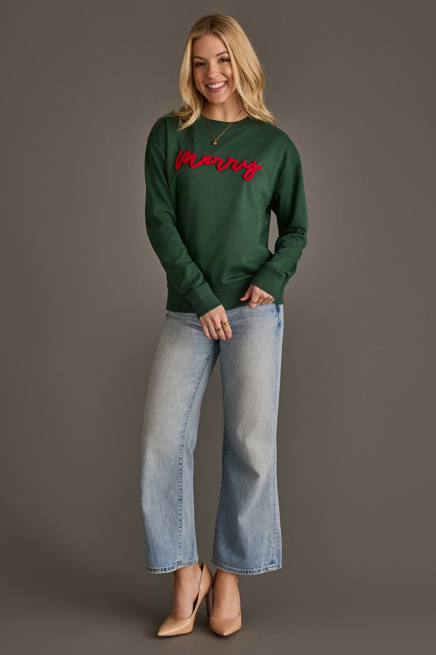 A person wearing a Dark Green Merry Sweatshirt with merry embroidered in red script across the chest, paired with light-wash wide-leg jeans and tan high heels.