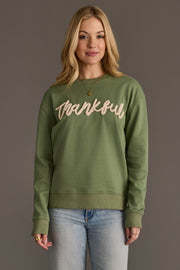 A woman with long blonde hair wears the Panache Apparel Collection’s Sage Green Thankful Sweatshirt paired with light blue jeans, standing against a plain gray background.