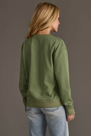 A person with long blonde hair, seen from behind, wears a Panache Apparel Collection Sage Green Thankful Sweatshirt and light blue jeans, standing against a plain gray background.