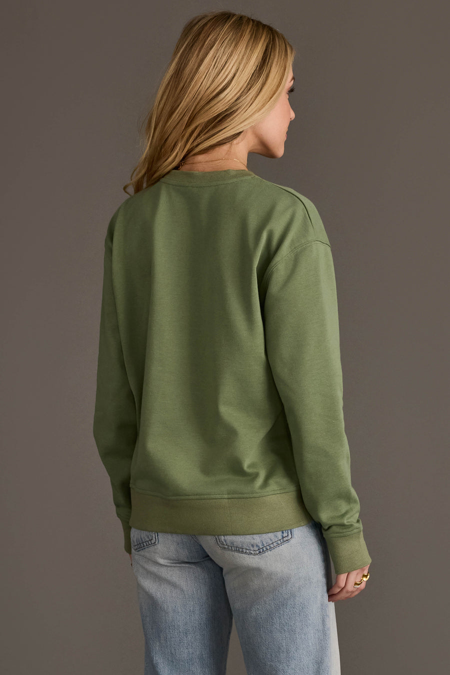 A person with long blonde hair, seen from behind, wears a Panache Apparel Collection Sage Green Thankful Sweatshirt and light blue jeans, standing against a plain gray background.