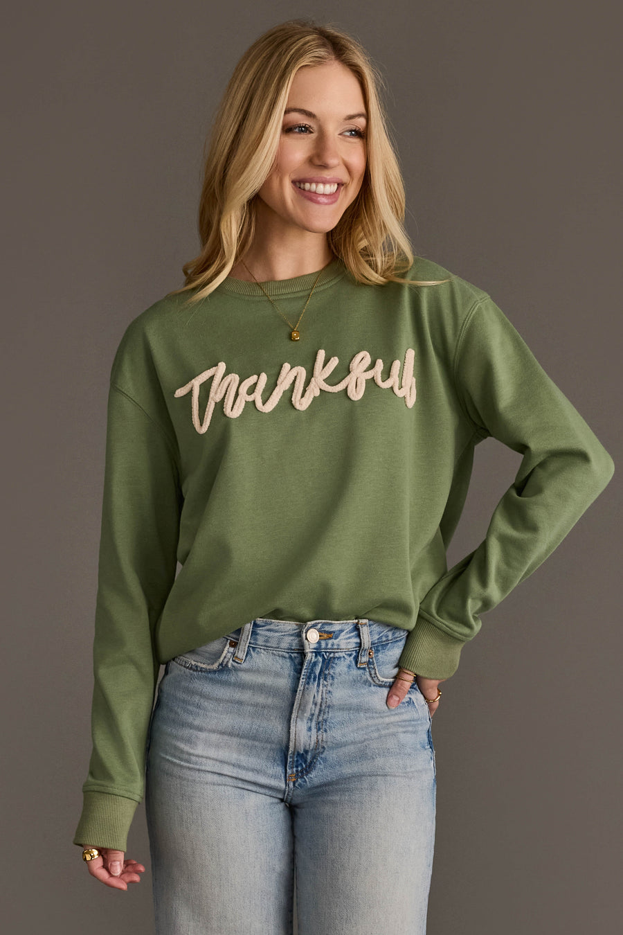 A smiling woman with long blonde hair wears the Panache Apparel Collection Sage Green Thankful Sweatshirt and light blue jeans, standing against a plain gray background.