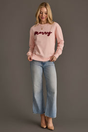 A woman wearing a Light Pink Merry Sweatshirt with dark red script and light-wash blue jeans.