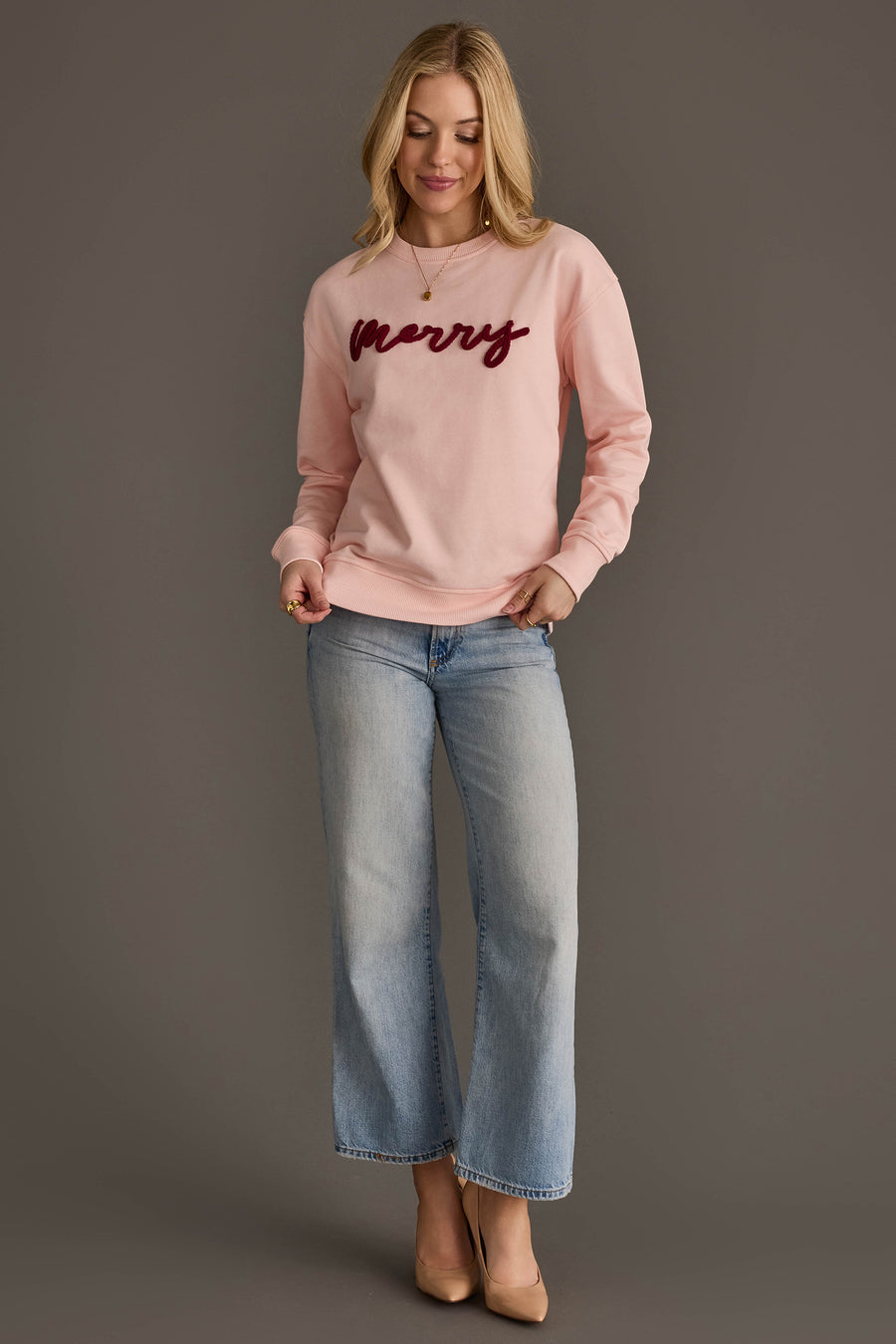 A woman wearing a Light Pink Merry Sweatshirt with dark red script and light-wash blue jeans.
