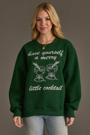 A person wears a Green Merry Little Cocktail Embroidered Sweatshirt featuring white line-art embroidery of two cocktails and the text have yourself a merry little cocktail.