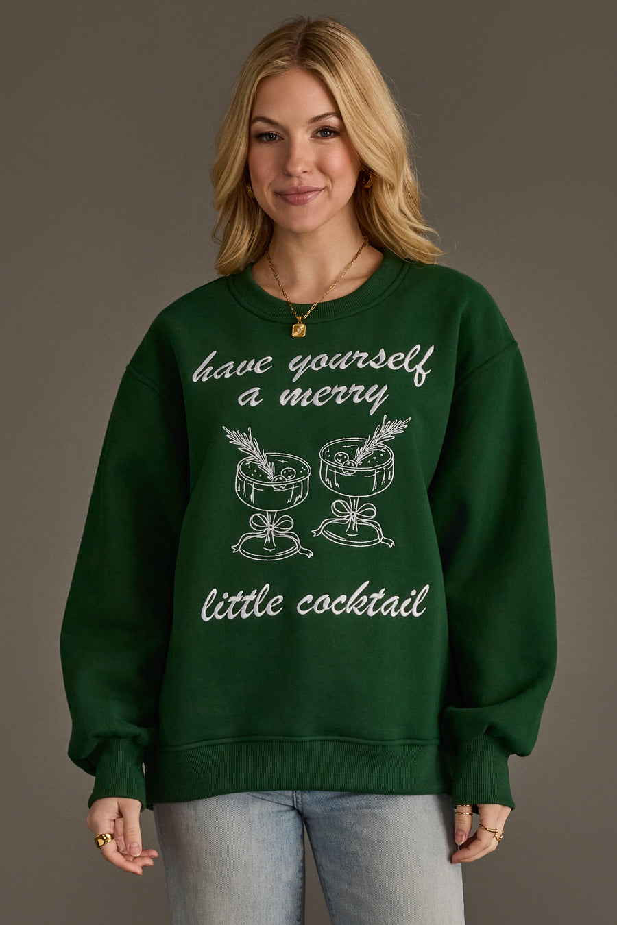 A person wears a Green Merry Little Cocktail Embroidered Sweatshirt featuring white line-art embroidery of two cocktails and the text have yourself a merry little cocktail.