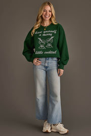 A smiling woman wears a green Merry Little Cocktail Embroidered Sweatshirt featuring an embroidered design of two cocktail glasses and the text have yourself a merry little cocktail.