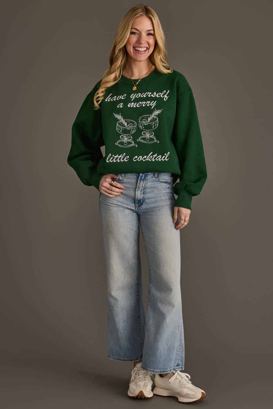 A smiling woman wears a green Merry Little Cocktail Embroidered Sweatshirt featuring an embroidered design of two cocktail glasses and the text have yourself a merry little cocktail.