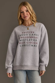 A person wearing a grey Panache Apparel Collection Christmas List Embroidered Sweatshirt featuring an oversized crewneck fit and a list of festive words embroidered in red and green text.