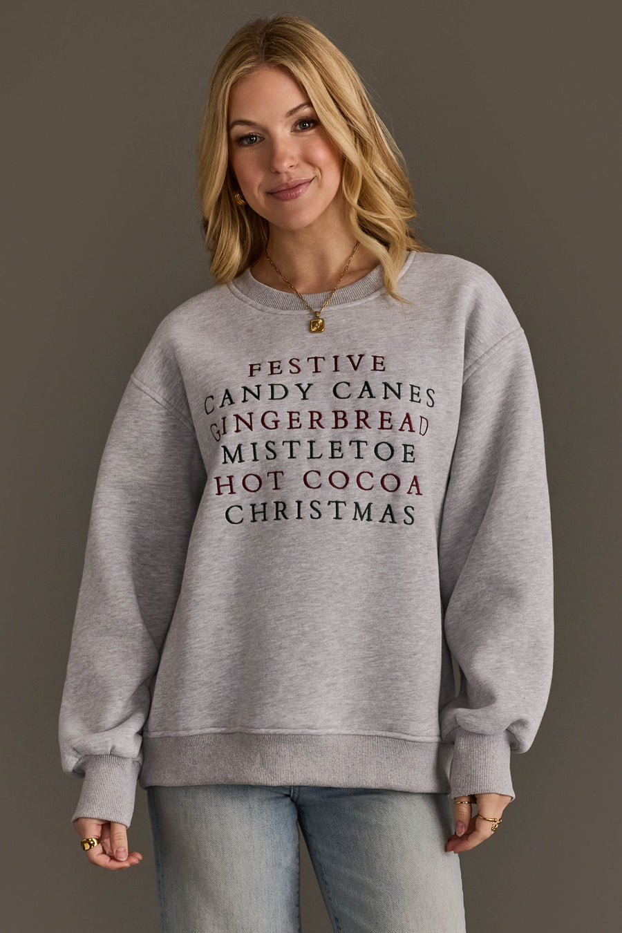A person wearing a grey Panache Apparel Collection Christmas List Embroidered Sweatshirt featuring an oversized crewneck fit and a list of festive words embroidered in red and green text.